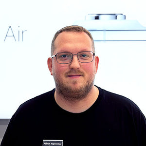 Ein Mann mit kurzen Haaren, einem Bart und einer Brille lächelt leicht in die Kamera. Er trägt ein schwarzes Hemd mit einem Namensschild. Ein weißer Hintergrund zeigt einen Teil des Wortes Air und einen metallischen Gegenstand.