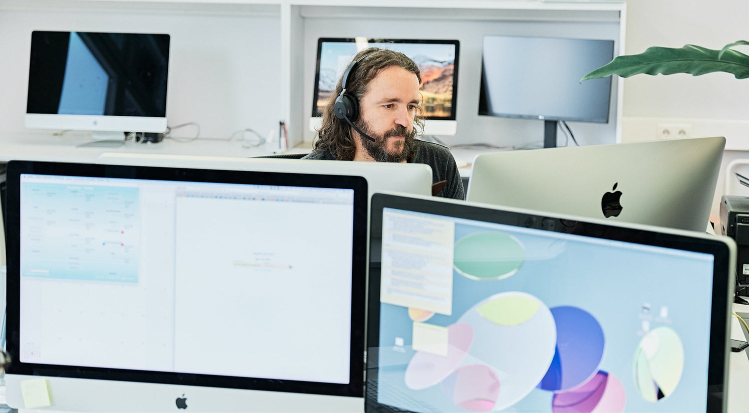 Ein Mann mit einem Headset arbeitet an einem Schreibtisch mit mehreren Apple-Computermonitoren in einem hellen, modernen Büro. Im Hintergrund sind weitere Computer und eine Pflanze zu sehen.