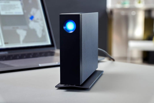 Eine schwarze LaCie d2 Professional 8TB USB-C-Desktop-Festplatte mit einem leuchtenden blauen Ring liegt auf einem Schreibtisch vor einem unscharfen offenen Laptop und bietet zuverlässigen Speicherplatz.