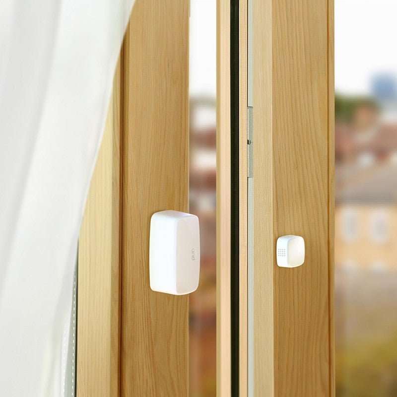 Eine Nahaufnahme von zwei Elgato Eve Door & Window, Smarter Kontaktsensor-Geräten, die an einem hölzernen Fensterrahmen befestigt sind, mit transparenten Vorhängen auf der linken Seite und einer verschwommenen Stadtlandschaft draußen.