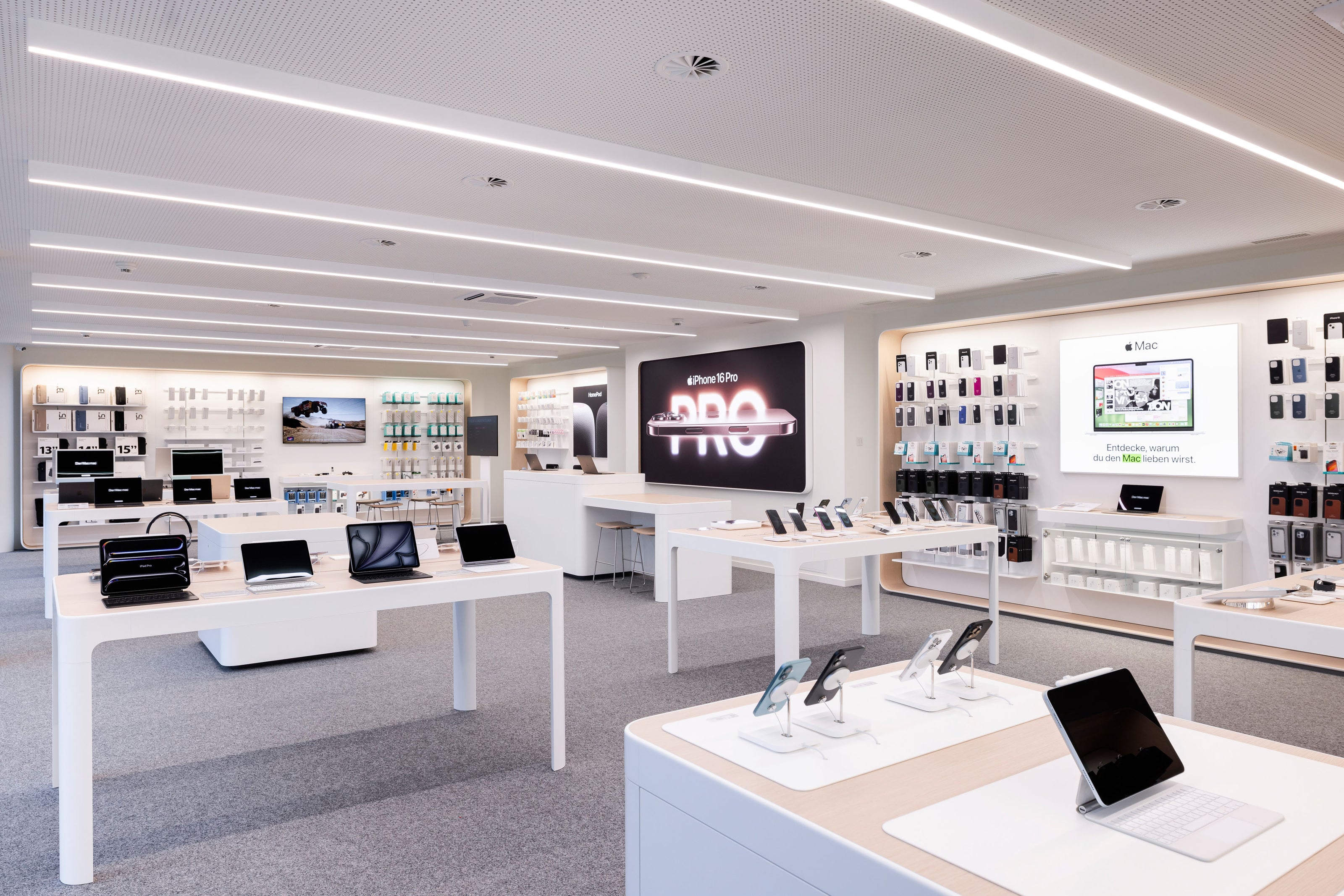 Ein modernes Elektronikgeschäft mit weißen Tischen, auf denen Apple-Laptops, -Tablets und -Smartphones ausgestellt sind. In den Regalen an den Wänden findet sich technisches Zubehör. Helle Beleuchtung und ein sauberes, minimalistisches Design sorgen für ein schlankes, organisiertes Erscheinungsbild.