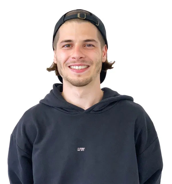 Ein junger Mann mit schulterlangen braunen Haaren, der eine schwarze Mütze und einen schwarzen Kapuzenpulli mit der Aufschrift LFDY trägt, lächelt, während er vor einem schlichten weißen Hintergrund steht.