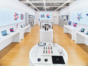 Ein heller, moderner Elektronikladen mit Holzböden, weißen Wänden und Auslagen mit Smartwatches, Tablets und Laptops. An den Wänden hängt Kunst und die Geräte sind ordentlich auf runden und rechteckigen Tischen angeordnet.