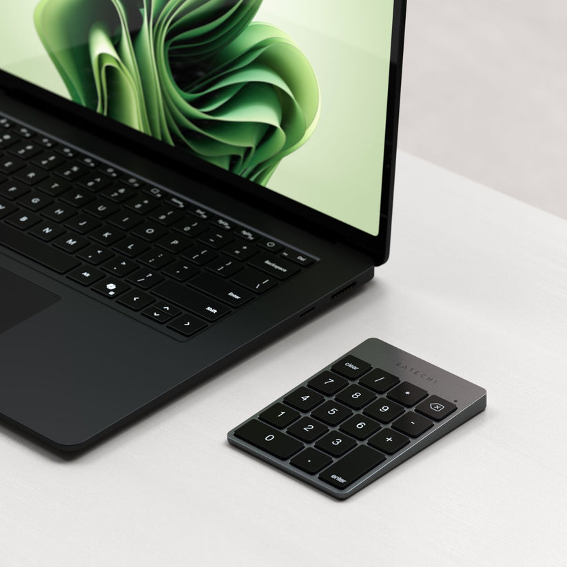 A sleek laptop with a green abstract wallpaper sits on a white desk next to the Satechi Slim Wireless Keypad Space Gray by Satechi, offering convenient number entry with its stylish and compact design.