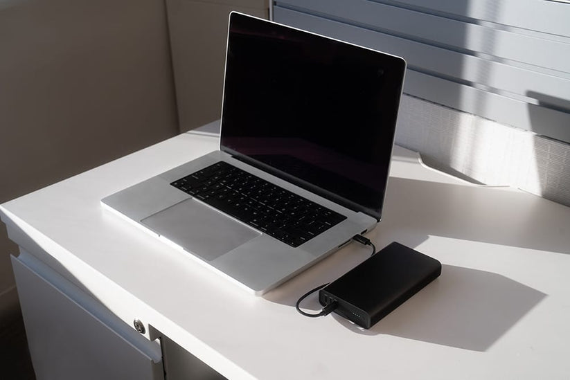 Auf einem weißen Schreibtisch in hellem Sonnenlicht, mit geschlossenen Jalousien im Hintergrund, ist ein Laptop an eine schwarze externe Festplatte und eine elegante „mophie Powerstation Ultra 25K – Schwarz“ von mophie angeschlossen, die dank ihrer beeindruckenden Kapazität von 25.000 mAh schnelles Laden und zuverlässige Backup-Möglichkeiten bietet.