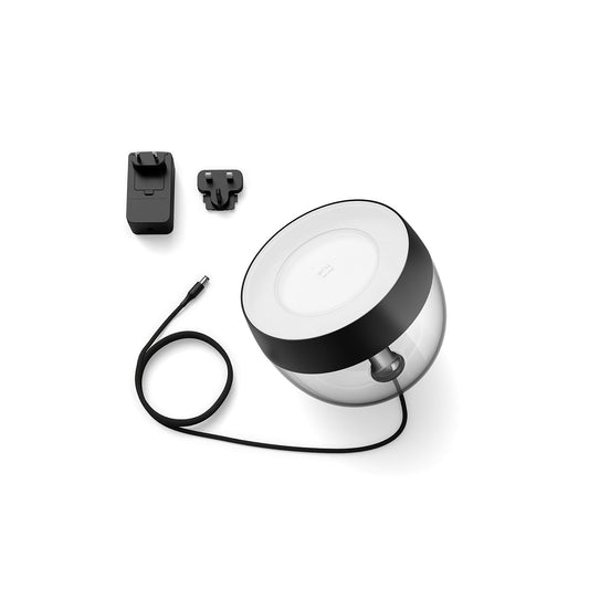 Die Philips Hue Iris Tischleuchte, schwarz von Philips ist eine transparente, runde LED-Lampe mit schwarzer Umrandung und Netzkabel, abgebildet mit einem schwarzen Wandadapter und Steckeraufsatz auf weiß. Die App-Steuerung ermöglicht eine bequeme Fernsteuerung des Lichts.
