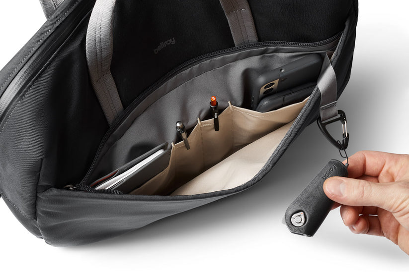Eine Hand hält einen schwarzen Schlüsselhalter, der an der Bellroy Via Work Bag (Tech Briefcase) in der Farbe „Slate“ befestigt ist. Im Inneren der Bellroy-Tasche sind Gegenstände wie Stifte, ein Handy und ein Notizbuch übersichtlich in beigen Fächern verstaut, was das praktische Design dieser wasserfesten Laptoptasche unterstreicht.