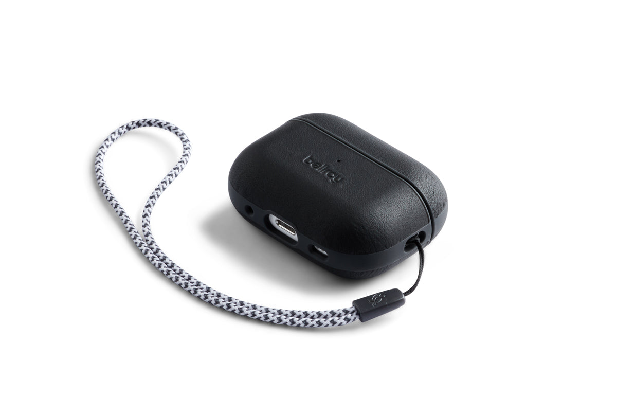 Eine Bellroy AirPods Jacket Pro (2. Generation) in Schwarz, die sich durch eine rechteckige Form mit einer strukturierten Oberfläche aus Premium-Leder und einem geflochtenen schwarz-weißen Handgelenkband auszeichnet, dargestellt vor einem weißen Hintergrund.
