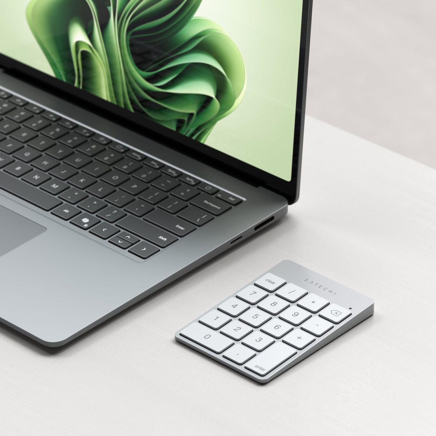 A laptop with a green abstract wallpaper sits on a light desk next to the Satechi Slim Wireless Keypad Silver, a sleek silver kabellose Zifferntastatur from Satechi clearly labeled with the brand name.