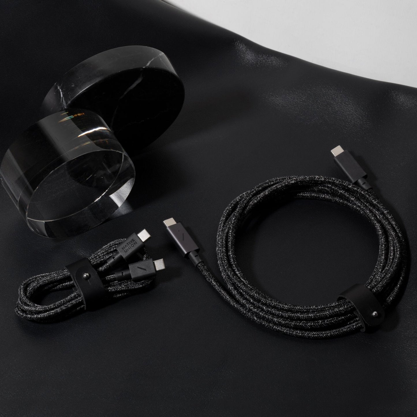Zwei „Native Union Belt Cable“ USB-C-zu-USB-C-Ladekabel (1,2 m, „Cosmos“) von Native Union, beide in Schwarz mit geflochtenem Design, sind ordentlich auf einer schwarzen Fläche neben zwei zylindrischen Objekten aus klarem Acryl aufgerollt.