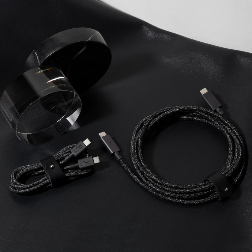 Zwei „Native Union Belt Cable“ USB-C-zu-USB-C-Ladekabel (1,2 m, „Cosmos“) von Native Union, beide in Schwarz mit geflochtenem Design, sind ordentlich auf einer schwarzen Fläche neben zwei zylindrischen Objekten aus klarem Acryl aufgerollt.