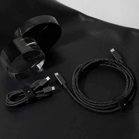 Native Union markasına ait iki adet Native Union Belt Cable USB-C - USB-C 1,2 m Cosmos şarj kablosu, her ikisi de siyah renkli ve örgülü tasarıma sahip olup, iki adet şeffaf akrilik silindirik nesnenin yanında siyah bir yüzey üzerinde düzgün bir şekilde sarılmış durumda.