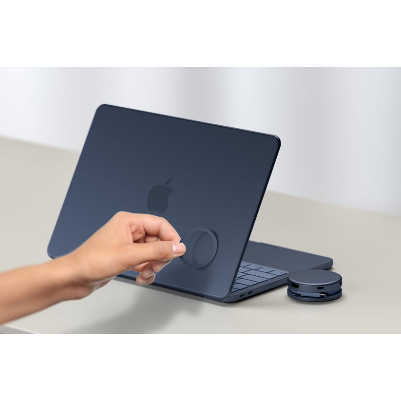 Eine Hand bringt ein rundes Klebepflaster auf der Rückseite eines geschlossenen, dunkelblauen Apple-Laptops auf einer hellen Oberfläche an, wobei der Satechi OntheGo 5-in-1-Multiport-Adapter (Indigo) von Satechi neben dem Laptop liegt.