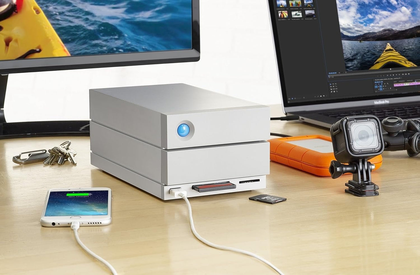 A LaCie 2big Dock Thunderbolt 3 Hardware RAID, 8TB sits on a desk, its silver design featuring a glowing blue button. It’s connected for iPad storage expansion, with a GoPro camera, orange portable drive, keys, and kayak photos on two monitors nearby.