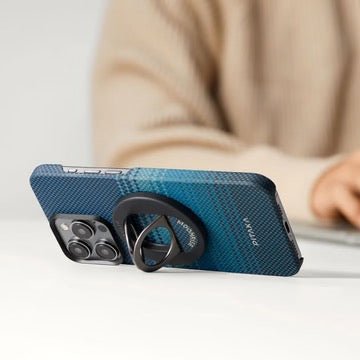 Eine blaue, gemusterte Smartphone-Hülle, an deren Rückseite ein runder, klappbarer PITAKA MagSafe Phone Ring Moonrise befestigt ist, liegt auf einer weißen Oberfläche.
