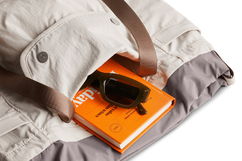 Eine Bellroy Cinch Tote in der Farbe „Oat“ mit braunen Trägern und einer gefalteten, wasserabweisenden leichten Jacke im Inneren, auf der ein orangefarbenes Buch und eine dunkle Sonnenbrille liegen, vor einem weißen Hintergrund.