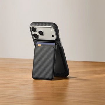 Eine schwarze Pitaka Magnetic Stand Woven Wallet in Schwarz/Grau (Twill), die an der Rückseite eines Smartphones befestigt ist und ausgeklappt auf einer Holzoberfläche als Ständer dient.