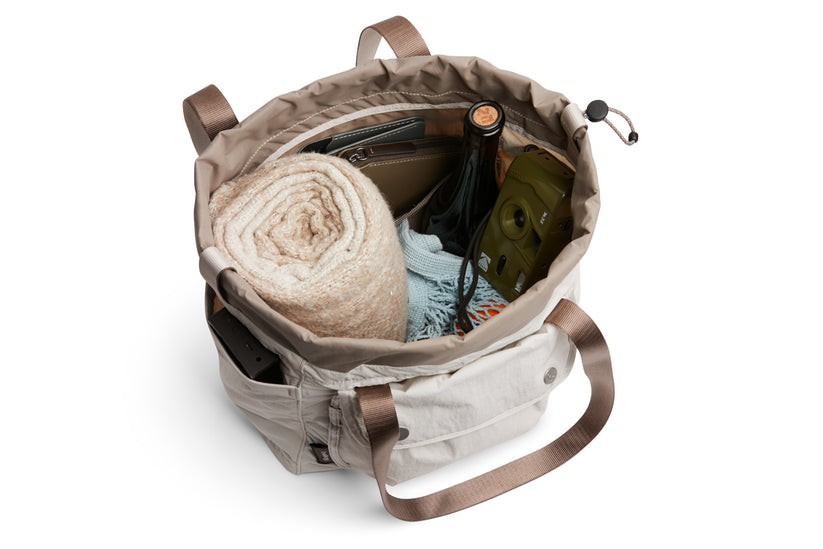 Die Bellroy Cinch Tote – Oat von Bellroy ist eine beigefarbene Tragetasche mit braunen Henkeln und einem verstellbaren Kordelzugverschluss. Sie bietet ausreichend Platz für all Ihre wichtigsten Utensilien, wie zum Beispiel eine zusammengerollte Decke, eine Flasche, ein Brillenetui, eine Geldbörse und eine Kamera. Die große Tasche ist zudem wasserabweisend.
