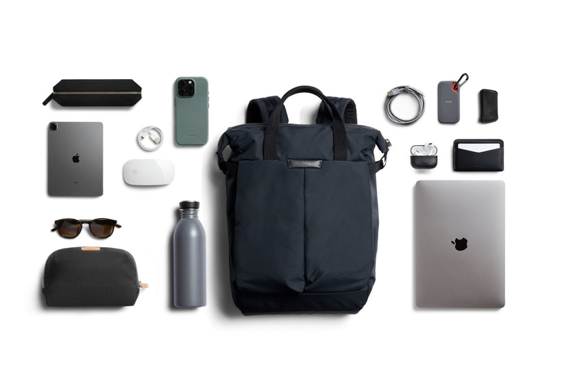 Im Mittelpunkt steht ein kompakter Rucksack, der Bellroy Tokyo Totepack Compact in Navy – umgeben von Elektronik, Zubehör und wichtigen Utensilien wie Laptop, Tablet, Handy, Sonnenbrille, Wasserflasche, Maus, Kabeln, Ladegerät, AirPods, Geldbörse und einem kleinen Etui. Alles ist ordentlich auf weißem Hintergrund angeordnet. Das durchdachte Laptopfach und das wasserabweisende Material machen diese Tasche von Bellroy besonders praktisch für den Alltag.