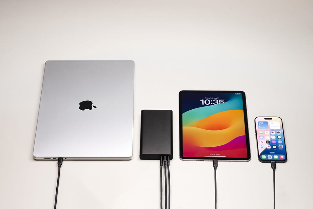 Kapalı bir MacBook, bir taşınabilir şarj cihazı, renkli ekranlı bir iPad ve bir iPhone, beyaz bir yüzey üzerinde yan yana şarj oluyor; her biri siyah bir şarj kablosuyla bağlı – tüm cihazlar, mophie’nin “mophie Powerstation Pro 20K – Siyah” ürünü tarafından verimli bir şekilde güçle besleniyor.