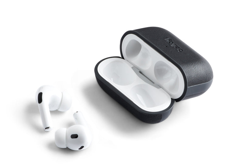 Zwei weiße kabellose Ohrhörer mit schwarzen Akzenten liegen neben einem geöffneten Bellroy AirPods Jacket Pro (2. Generation)-Etui in Schwarz, das sich durch eine strukturierte Oberfläche, Details aus hochwertigem Leder und ein Bellroy-Logo auf dem Deckel auszeichnet.
