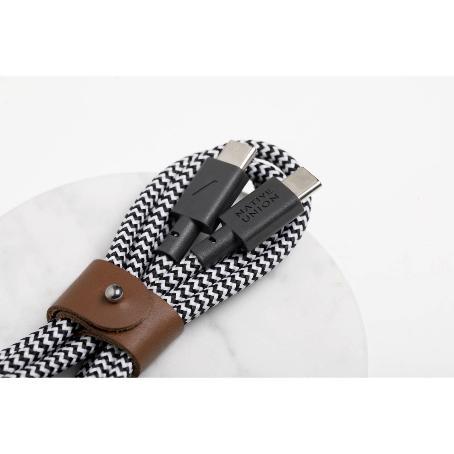 Ein Native Union Belt-Cable USB-C-zu-USB-C-Kabel (1,2 m) in der Farbvariante „Zebra“ mit schwarz-weißem Flechtdesign und einem braunen Lederband ist auf einer weißen Unterlage aufgerollt. Auf den Steckern ist der Markenname „Native Union“ zu sehen.