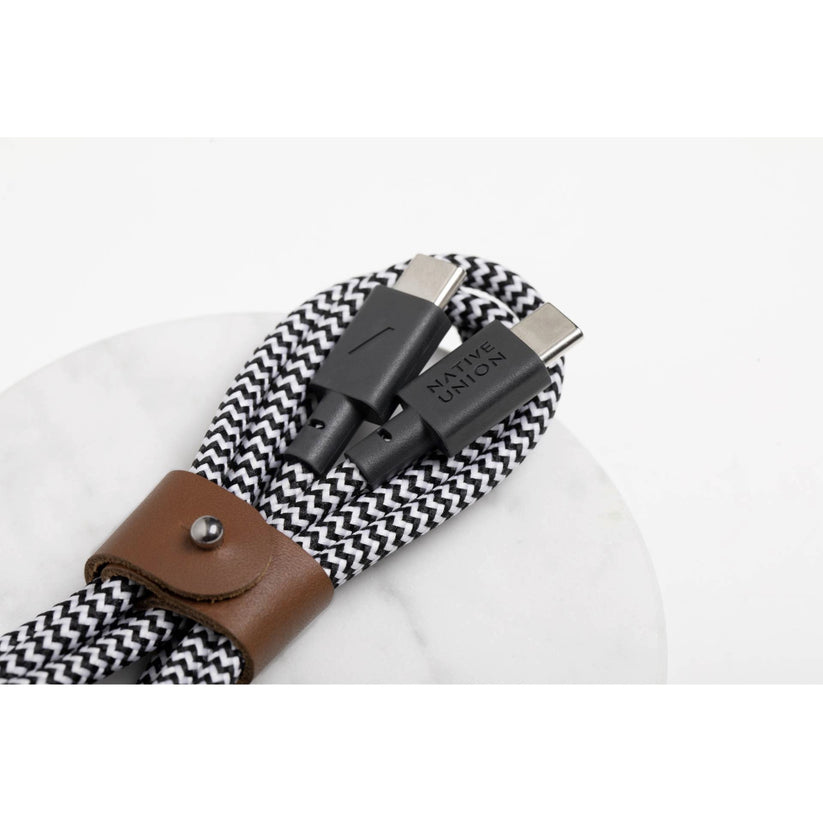 Ein Native Union Belt-Cable USB-C-zu-USB-C-Kabel (1,2 m) in der Farbvariante „Zebra“ mit schwarz-weißem Flechtdesign und einem braunen Lederband ist auf einer weißen Unterlage aufgerollt. Auf den Steckern ist der Markenname „Native Union“ zu sehen.