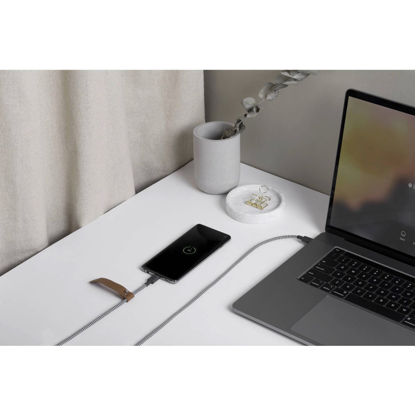 Ein „Native Union Belt-Cable“ USB-C-zu-USB-C-Kabel (1,2 m, Zebra-Muster) lädt ein Smartphone neben einem Laptop auf einem weißen Schreibtisch auf; daneben steht eine kleine Schale mit Schmuck und ein grüner Zweig.