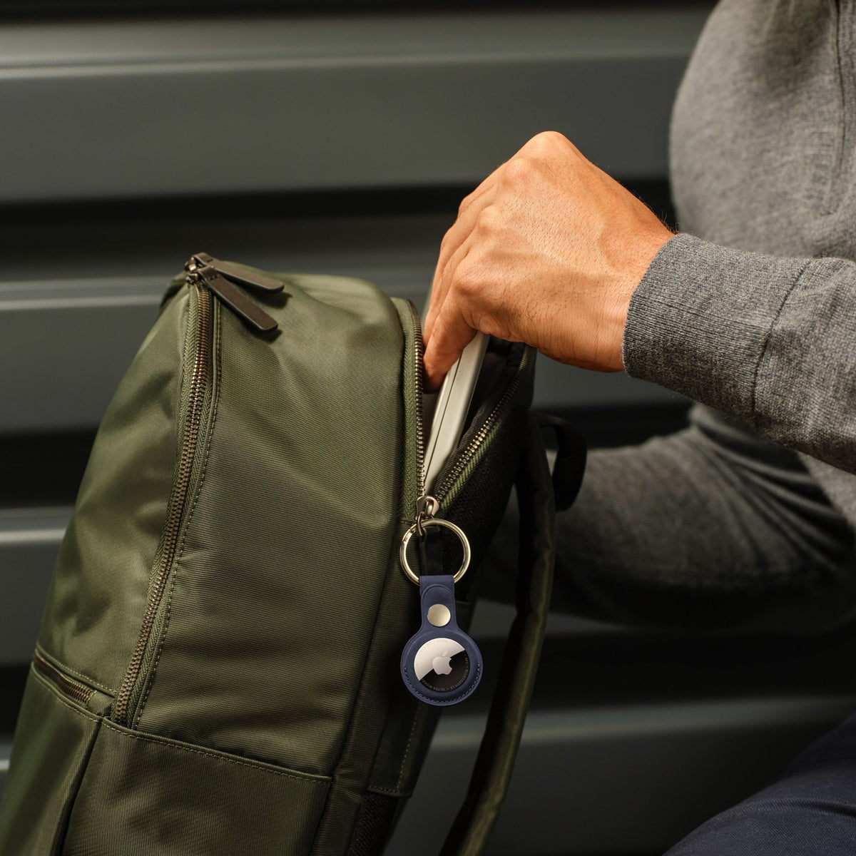 A person tucking an Apple AirTag, 1er-Pack (2026) in a navy blue key ring holder into the zippered compartment of a green backpack.