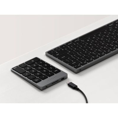 Ein Satechi Slim Wireless Keypad in Space Gray und eine dazu passende Tastaturauflage liegen auf einer weißen Oberfläche. Daneben liegt ein abnehmbares USB-C-Kabel, das jederzeit an das kabellose Satechi-Keypad angeschlossen werden kann.