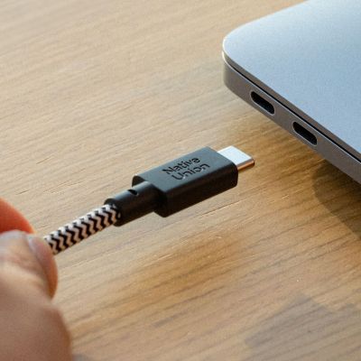 Eine Hand hält ein Native Union Belt Cable XL USB-C-zu-USB-C-Kabel (3 m, Zebra-Muster) in der Nähe eines Laptops mit zwei USB-C-Anschlüssen, bereit zum Anschließen. Das geflochtene Kabel, auf dessen Stecker „Native Union“ zu lesen ist, ist auf Langlebigkeit ausgelegt – und damit die perfekte 3-Meter-Lösung zum Laden und Synchronisieren.