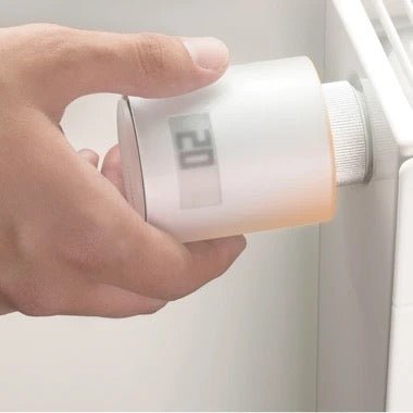 Eine Hand verstellt den Drehknopf eines Netatmo Smarte Heizkörperthermostate Starterpaketes an einem Heizkörper, dessen Display 20-demonstrating eine intelligente Beheizungslösung von Netatmo für smarte Heizkörperthermostate anzeigt.