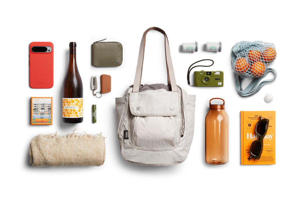 Die Bellroy Cinch Tote – Oat, eine weiße, wasserabweisende Tasche von Bellroy, ist auf einem weißen Hintergrund abgebildet, umgeben von einem Handy, einer Geldbörse, Schlüsseln, einer Flasche, einer Kamera, Orangen in einem Netzbeutel, einer Sonnenbrille, einem Buch, einer Strickdecke, einer Weinflasche und kleinen Gegenständen. Dank ihrer geräumigen Fächer eignet sie sich perfekt zum Ordnen Ihrer wichtigsten Dinge.