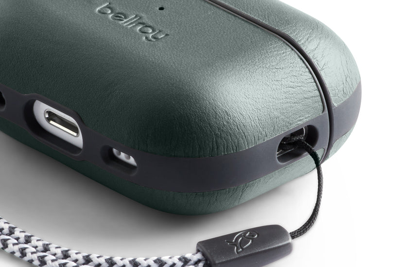 Eine Nahaufnahme der Bellroy AirPods Jacket Pro (2. Generation) – Everglade mit integriertem Trageband. Das Bild zeigt die strukturierte Oberfläche aus Premium-Leder, einen USB-C-Ladeanschluss sowie einen Ausschnitt des markanten schwarz-weißen Musters des Tragebands.