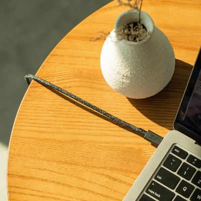 Eine Nahaufnahme eines Laptops auf einem Holztisch, an den das Native Union Belt Cable XL USB-C-zu-USB-C-Kabel (3 m, Farbe: Cosmos) angeschlossen ist; im Hintergrund steht eine strukturierte weiße Vase mit Trockenblumen. Sonnenlicht wirft sanfte Schatten über die Szene.