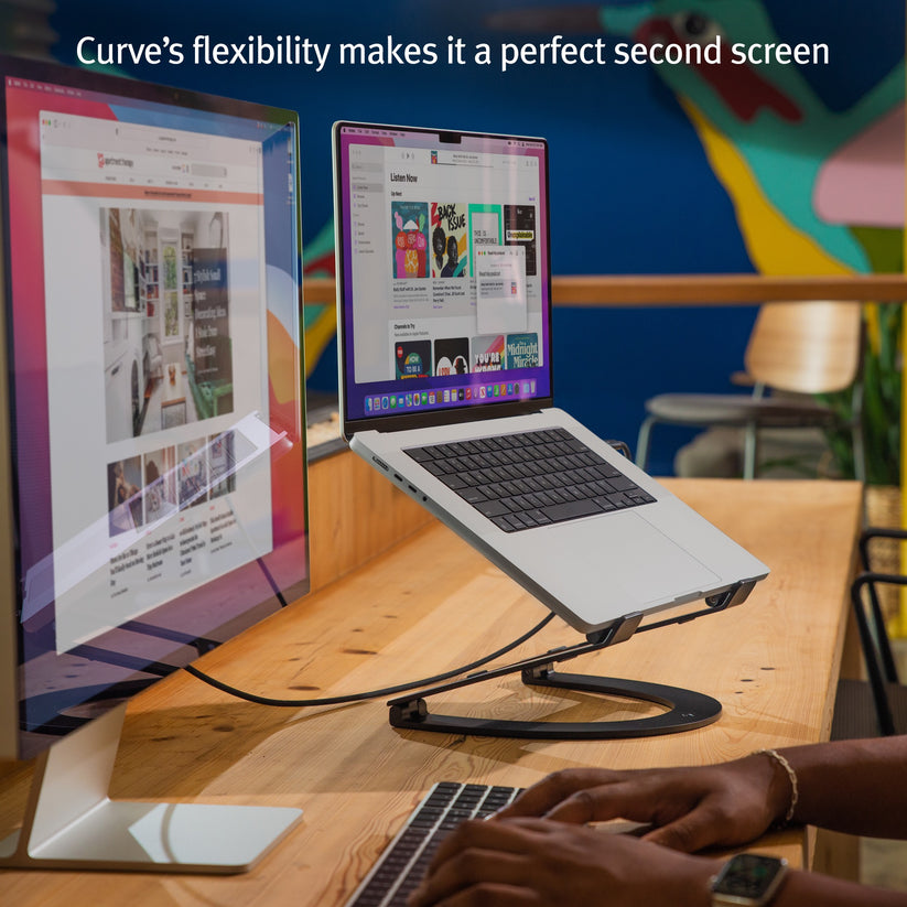 Ein weißer Twelve South Curve Flex Aluminium-Ständer für MacBook dient dazu, einen Laptop als zweiten Bildschirm neben einem Desktop-Monitor auf einem Holzschreibtisch zu befestigen, vor dem im Hintergrund farbenfrohe Kunstwerke zu sehen sind. Dank seiner flexiblen Kurven und der ergonomischen Arbeitshaltung ist der Twelve South Curve Flex das perfekte MacBook-Zubehör für jeden Arbeitsplatz.