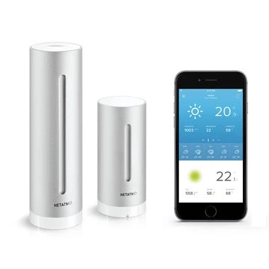 Zwei silberne Netatmo Smarte Wetter-Station Zylinder stehen neben einem Smartphone, das umfassende Klimadaten wie Temperatur, Luftfeuchtigkeit und Wettervorhersage auf dem Display anzeigt.