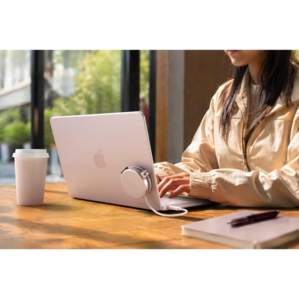 Eine Person in einer beigen Jacke tippt auf einem silbernen Apple-Laptop an einem Holztisch, neben dem eine Kaffeetasse, ein Notizbuch, ein Stift und kabellose Kopfhörer liegen. Neben dem Laptop steht der Satechi OntheGo 5-in-1-Multiport-Adapter (Blush) von Satechi, der nahtlose Konnektivität bietet, während Sonnenlicht durch die großen Fenster hereinströmt.