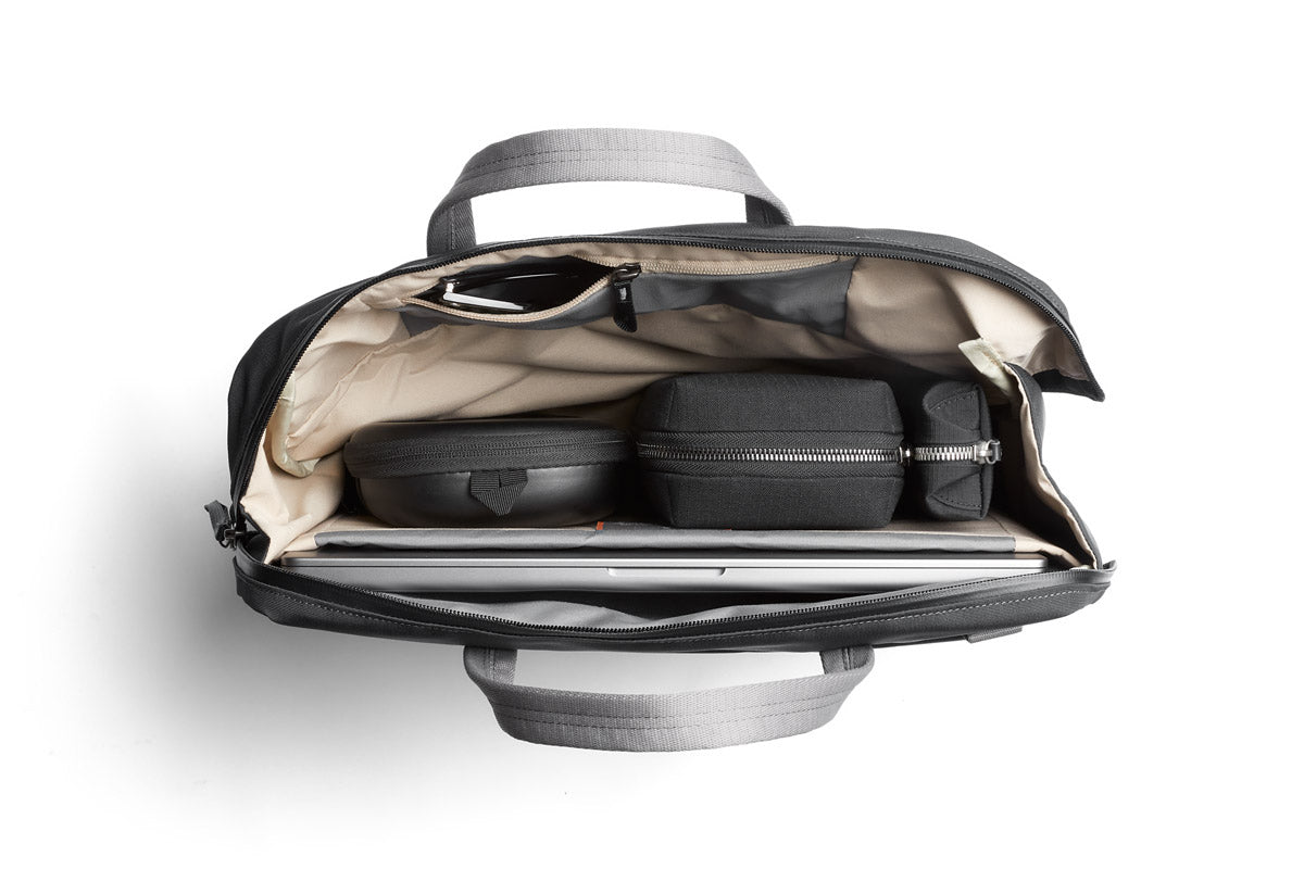 Ein Blick von oben auf eine geöffnete Bellroy Via Work Bag (Tech Briefcase) in der Farbe „Slate“ offenbart einen übersichtlich angeordneten Inhalt: eine Sonnenbrille, zwei schwarze Beutel mit Reißverschluss, ein kleines schwarzes Etui und ein Laptop, der in cremefarbenen Innentaschen verstaut ist. Diese wasserfeste Laptoptasche von Bellroy vereint Stil und Funktionalität für den täglichen Gebrauch.
