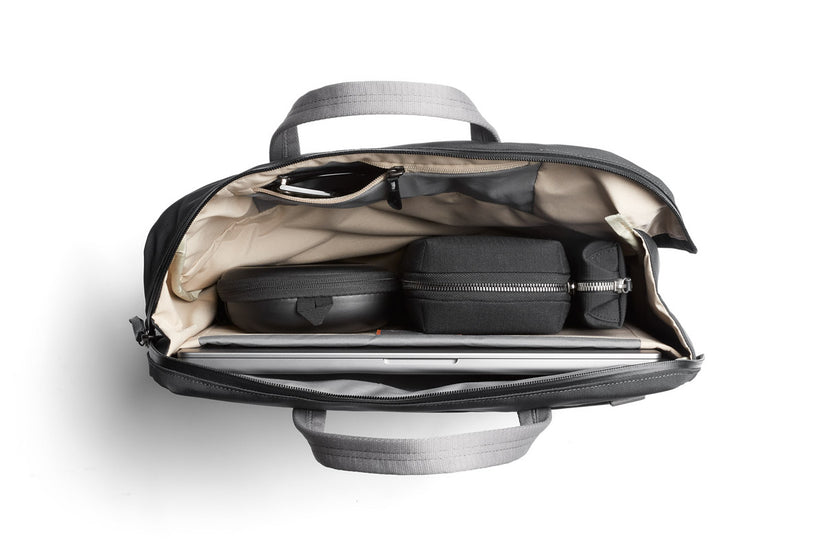 Ein Blick von oben auf eine geöffnete Bellroy Via Work Bag (Tech Briefcase) in der Farbe „Slate“ offenbart einen übersichtlich angeordneten Inhalt: eine Sonnenbrille, zwei schwarze Beutel mit Reißverschluss, ein kleines schwarzes Etui und ein Laptop, der in cremefarbenen Innentaschen verstaut ist. Diese wasserfeste Laptoptasche von Bellroy vereint Stil und Funktionalität für den täglichen Gebrauch.