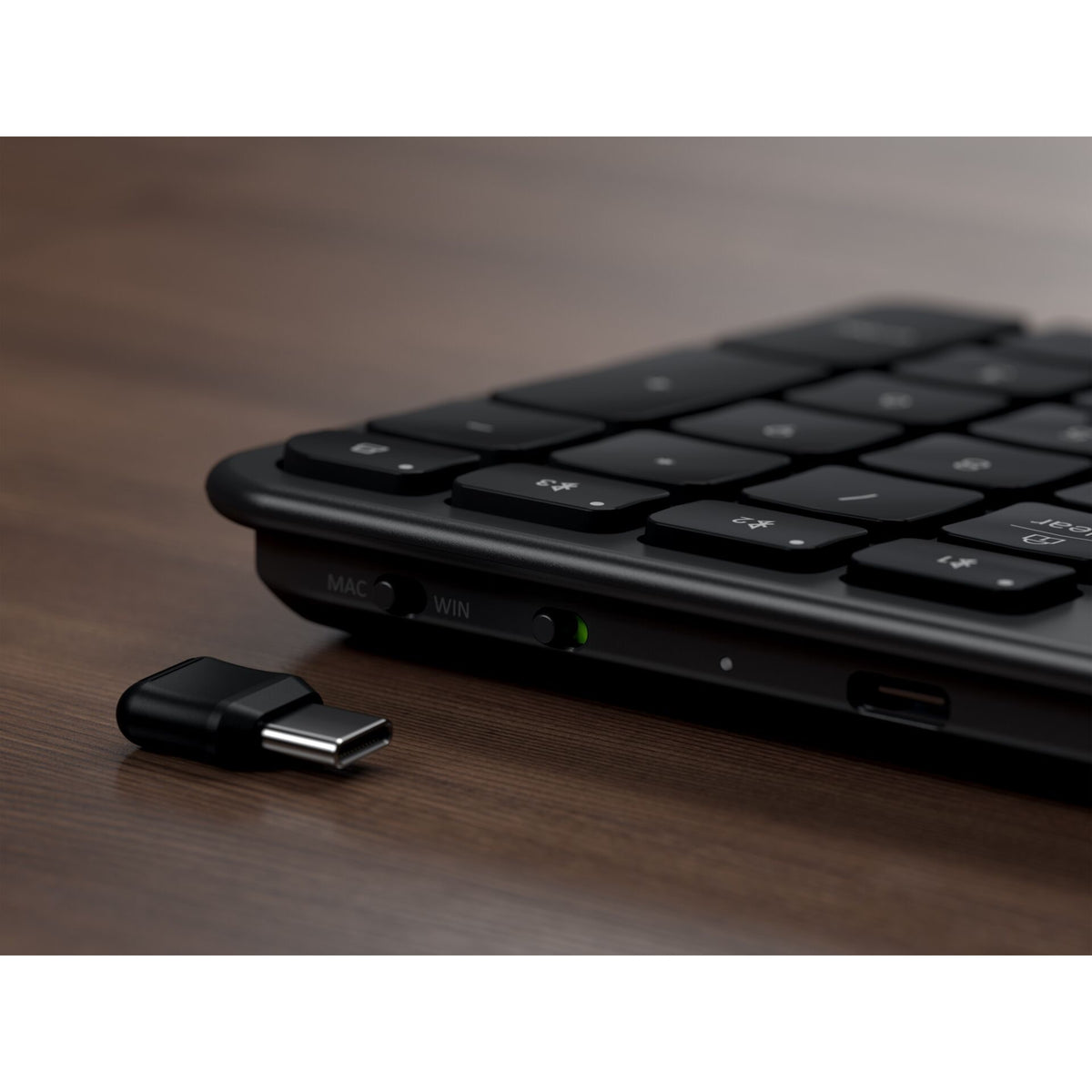 Eine Nahaufnahme einer Satechi Slim EX3 Wireless Keyboard Space Black DE mit deutschem Tastaturlayout auf einer Holzoberfläche; daneben ist ein USB-C-Dongle zu sehen, und an der Seite der Tastatur sind Schalter mit den Aufschriften „Mac“ und „Win“ zu erkennen.