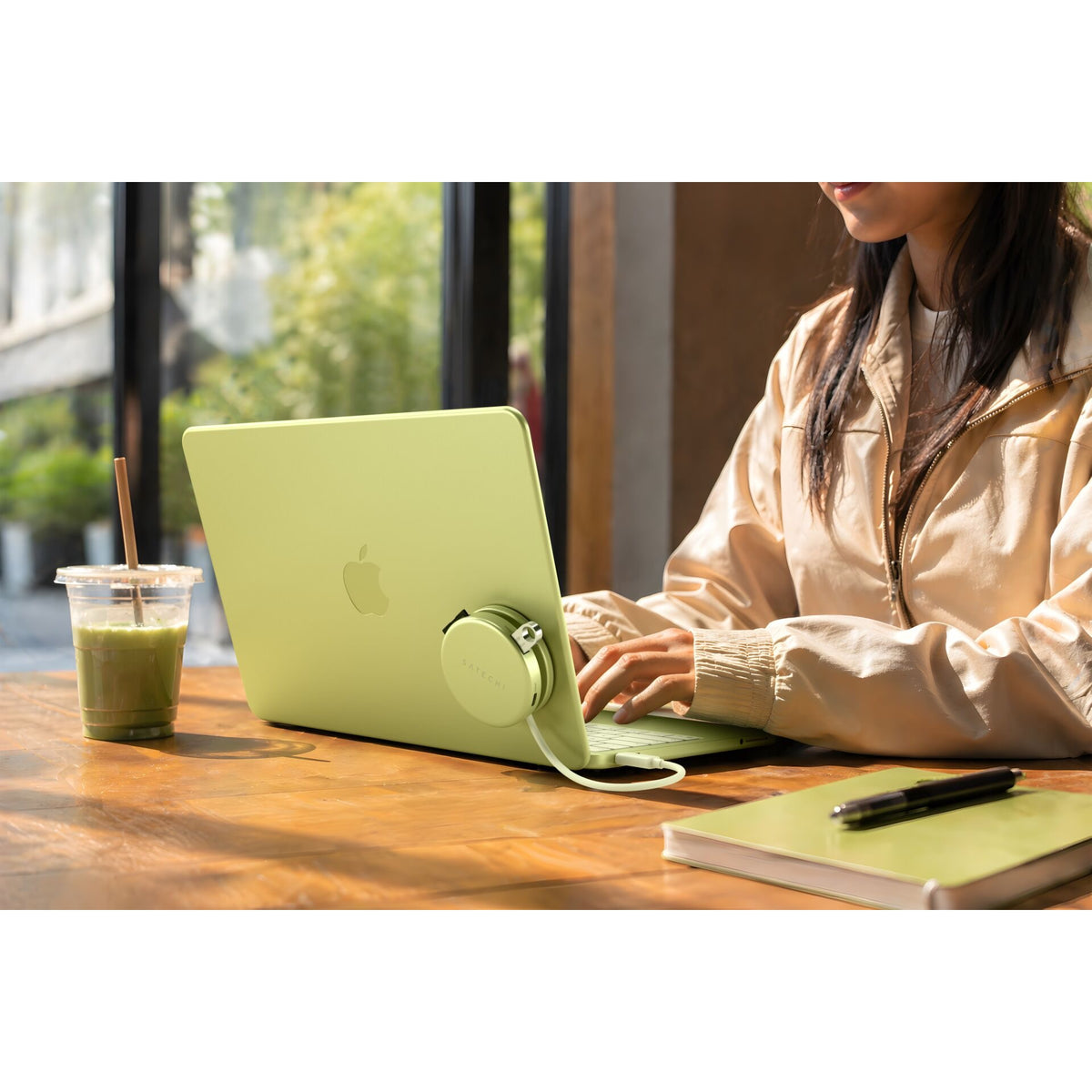 Eine Person tippt an einem hellgrünen Laptop, der auf einem Holztisch steht, umgeben von einem grünen Getränk, einem Notizbuch, einem Stift und Kopfhörern, während Sonnenlicht durch große Fenster hereinströmt. Ein Satechi OntheGo 5-in-1-Multiport-Adapter (Citrus) von Satechi ist an den Laptop angeschlossen und verbindet alle Geräte nahtlos miteinander, um effizientes Multitasking zu ermöglichen.