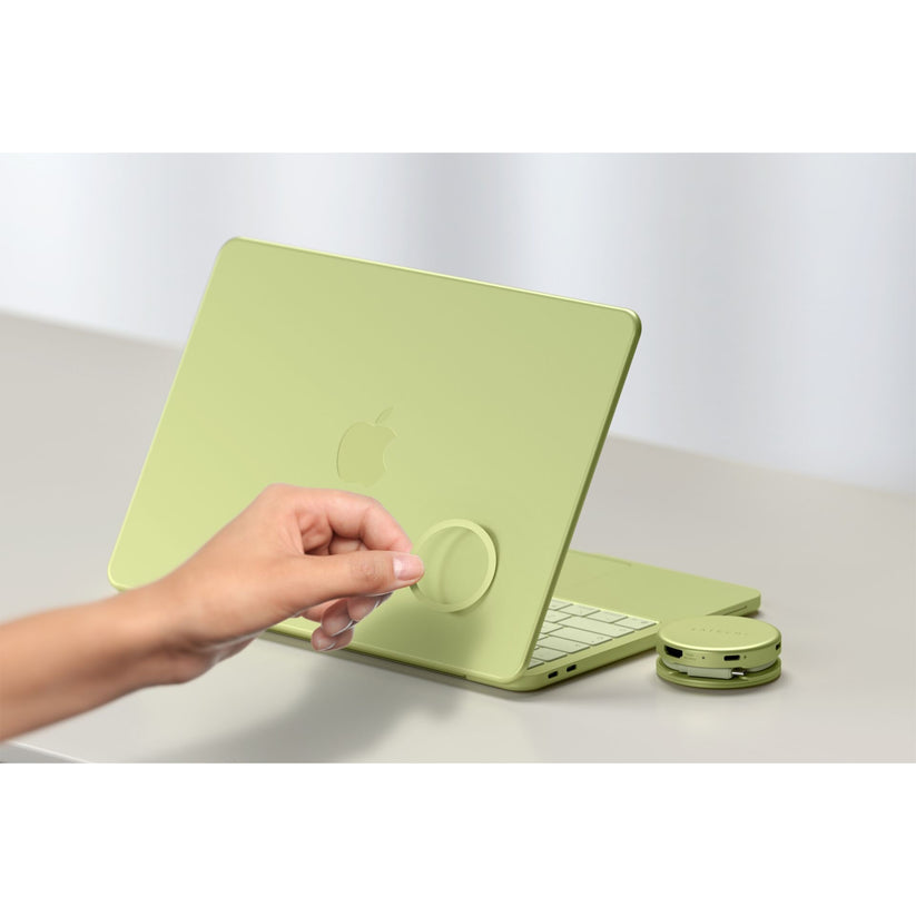 Eine Hand befestigt ein rundes, farblich passendes grünes Zubehörteil an der Rückseite eines grünen Apple-Laptops. Neben dem Laptop auf dem Tisch liegen eine farblich passende grüne kabellose Maus und der elegante Satechi OntheGo 5-in-1-Multiport-Adapter (Citrus) von Satechi, der sich perfekt zur Erweiterung Ihrer Anschlussmöglichkeiten eignet.