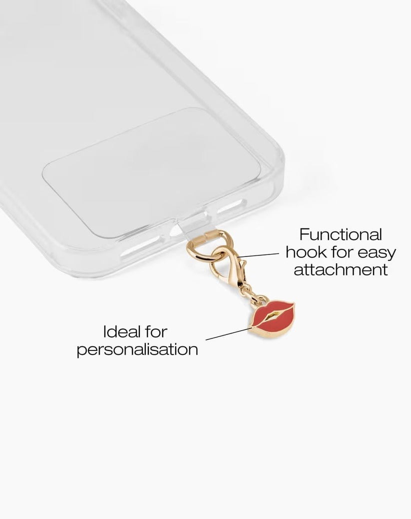 Eine transparente Handyhülle von Ideal of Sweden verfügt über einen goldenen Haken zum einfachen Befestigen, an dem der Ideal of Sweden-Anhänger „French Kiss“ – ein Handy-Charm in Form roter Lippen – befestigt werden kann. Der Text auf dem Bild hebt den praktischen Haken hervor und eignet sich somit perfekt zur individuellen Gestaltung.