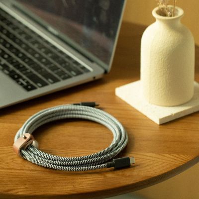 Ein spiralförmiges Native Union Belt Cable XL USB-C-zu-USB-C-Kabel (3 m, Zebra-Muster) liegt auf einem Holztisch neben einem geöffneten Laptop und einer strukturierten weißen Vase.