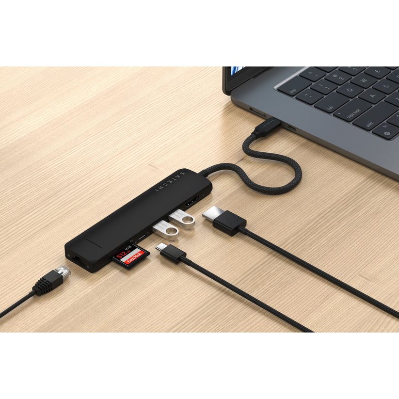Ein schwarzer Satechi USB-C 7-in-1 Slim Multiport-Adapter mit Ethernet ist an einen Laptop angeschlossen, wobei mehrere Kabel, USB-Sticks, SD-Karten und Adapter an den Hub angeschlossen sind – alles steht auf einer hellen Holzunterlage.