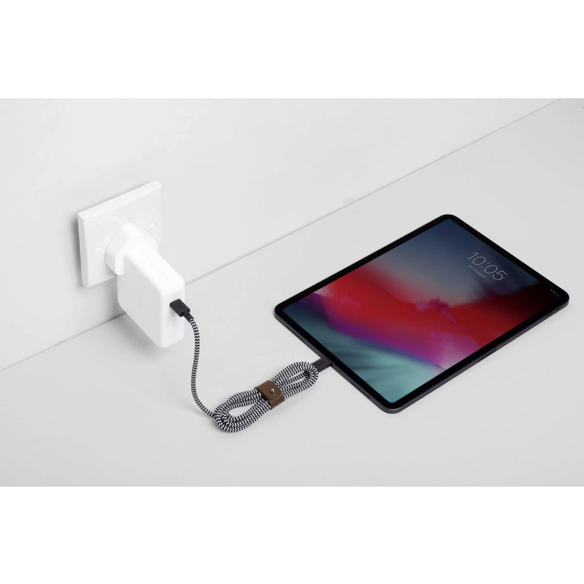 Ein Tablet liegt auf einer weißen Fläche neben einer Steckdose und ist über ein „Native Union Belt-Cable USB-C auf USB-C 1,2 m Zebra“ von Native Union angeschlossen; das Spiralkabel ist eingesteckt, während auf dem Bildschirm die Uhrzeit 10:05 angezeigt wird.