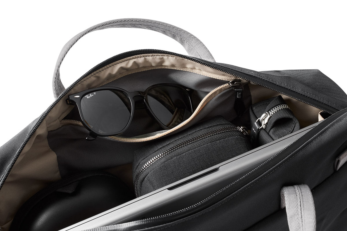 Eine Nahaufnahme einer geöffneten Bellroy Via Work Bag (Tech Briefcase) in der Farbe „Slate“ zeigt eine Sonnenbrille, ein kleines Etui mit Reißverschluss, einen aufgerollten Gegenstand und ein elektronisches Gerät, die ordentlich im Inneren angeordnet sind. Das hellbraune Innenfutter bildet einen Kontrast zu den grauen Tragegriffen der Tasche.