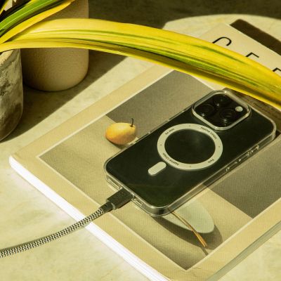 Ein Smartphone mit einem ringförmigen Design auf dem Gehäuse wird mit dem Native Union Belt Cable XL USB-C zu USB-C (3 m, Muster „Zebra“) aufgeladen, während es auf einer Zeitschrift liegt, auf deren Titelseite eine Birne abgebildet ist. Daneben sind außerdem ein gelbgrünes Blatt und ein Teil einer Topfpflanze zu sehen.