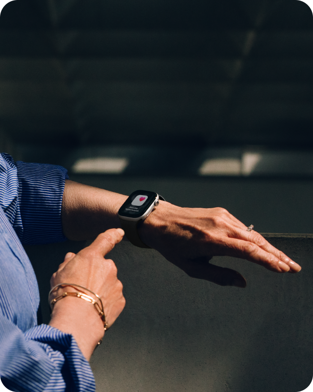 Eine Person, die ein blaues Hemd und goldene Armbänder trägt, interagiert in einem schwach beleuchteten Innenraum mit einer Smartwatch, die ein Herzsymbol anzeigt und möglicherweise die Herzfrequenz überwacht.